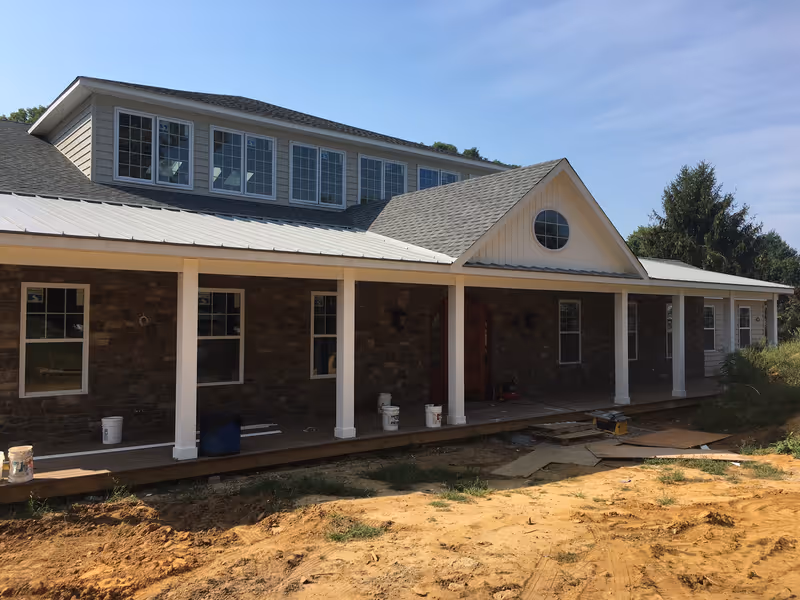Exterior view of a building under construction with a stone facade, white columns supporting a covered porch, multiple windows, and a gray shingled roof under a clear blue sky.