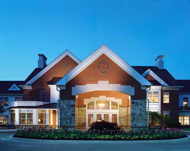 Front exterior view of a senior living facility building with brick and stone facade, large windows, and a well-maintained flower bed in front under a clear blue sky.