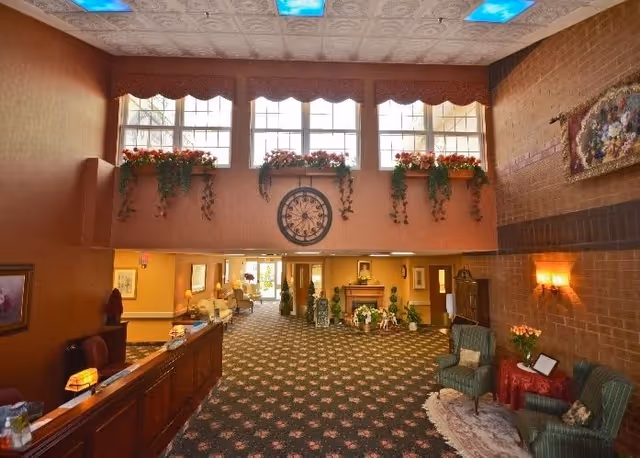 A spacious and warmly decorated nursing home lobby with a patterned carpet, a wooden reception desk on the left, and seating areas with armchairs and small tables on the right. Above the entrance, there are large windows with flower boxes and hanging plants, and a large decorative clock is mounted on the wall. The ceiling has decorative panels with blue light fixtures.