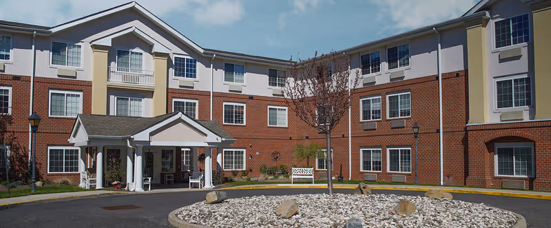Exterior view of a three-story senior living facility building with a brick and light-colored facade, multiple windows, a covered entrance with white columns, a small tree, and a circular rock garden in front.