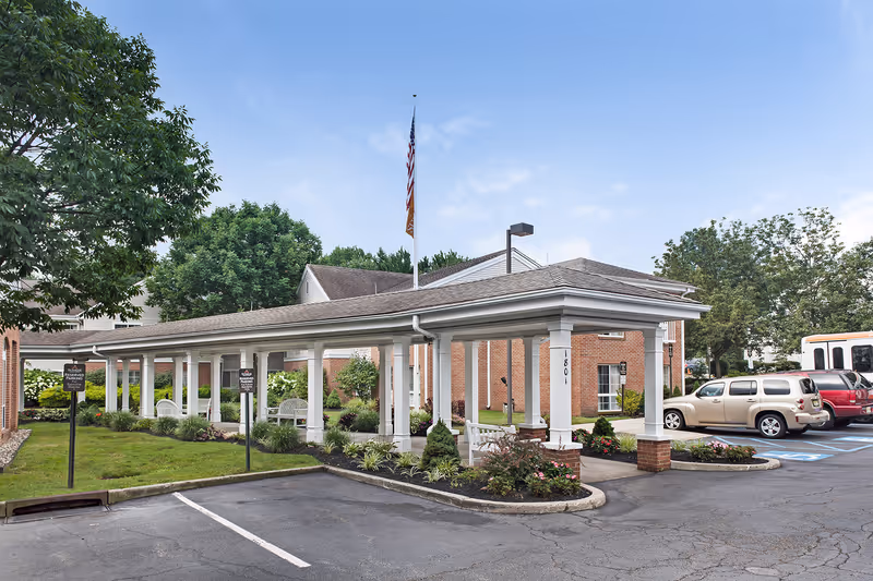 Exterior view of Brighton Gardens of Edison showing a covered walkway supported by white columns, surrounded by landscaped greenery and flower beds. Several parked cars and a flagpole with an American flag are visible under a clear blue sky.