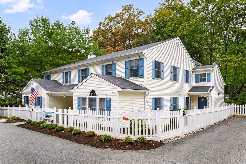 Exterior view of a two-story white building with blue shutters surrounded by a white picket fence. There is an American flag displayed near the entrance and a sign on the fence that reads 'Heroes Work Here'. The building is set against a backdrop of green trees under a partly cloudy sky.