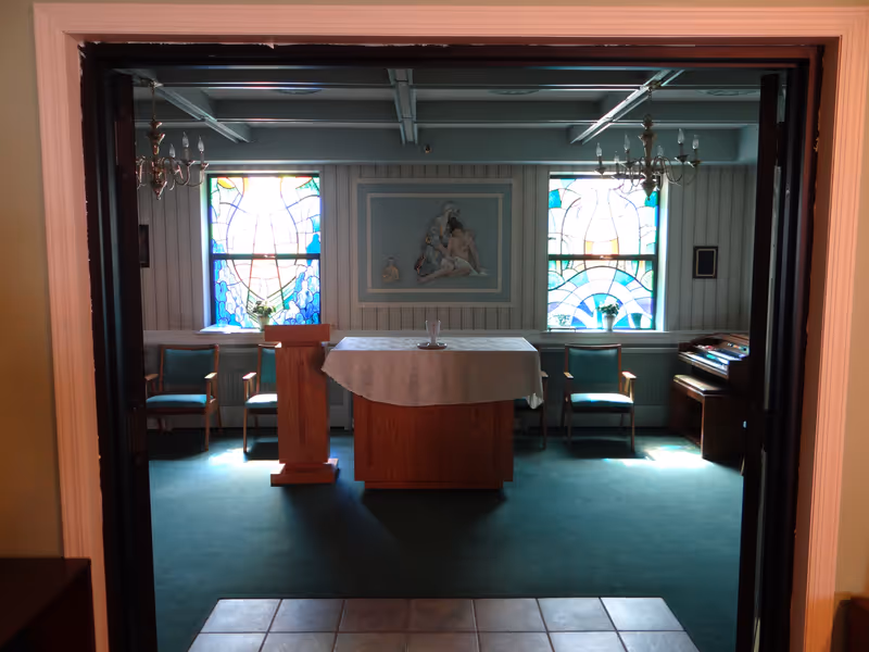 Interior view of a small chapel or prayer room with two stained glass windows, a wooden altar covered with a white cloth, a wooden lectern, four chairs, two chandeliers, and an organ on the right side. A religious painting is mounted on the wall between the windows.