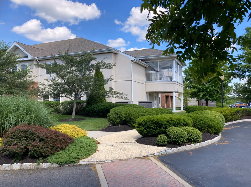 Exterior view of a two-story senior living facility building with beige walls and a gray roof, surrounded by well-maintained bushes, trees, and landscaped greenery under a partly cloudy blue sky.