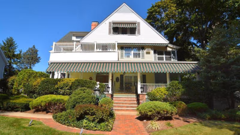 Front exterior view of a large white multi-story house with a green and white striped awning over the porch, surrounded by well-maintained bushes and trees under a clear blue sky.