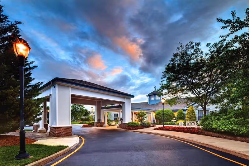 Covered porte-cochere entrance to a senior living building with a curved driveway, landscaped beds, lampposts and trees under a colorful evening sky.