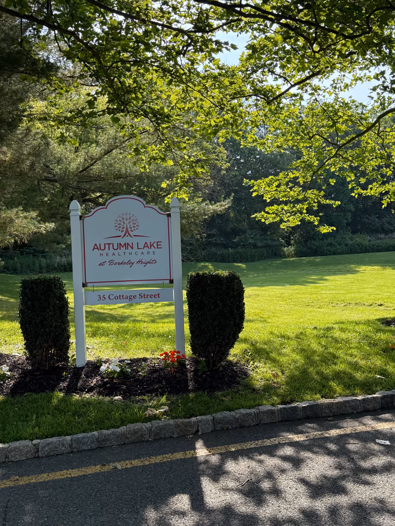 A white entrance sign reading 'Autumn Lake Healthcare at Berkeley Heights' on a grassy lawn surrounded by trees and shrubs.