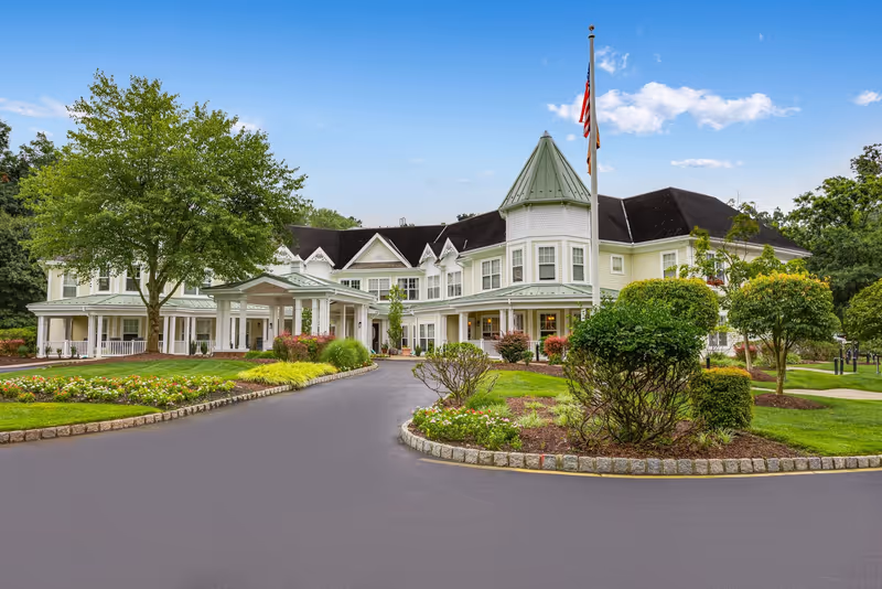 Front exterior view of a large senior living facility building with a covered entrance, manicured gardens, trees, and an American flag on a flagpole under a partly cloudy blue sky.