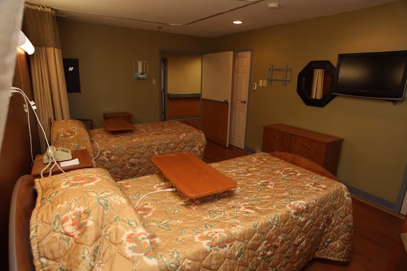 A room with two single beds covered in floral patterned bedspreads. Each bed has a wooden overbed table. There is a wooden dresser with a wall-mounted TV above it and a small octagonal mirror on the wall. The walls are painted green and the floor is wooden. A door is open, showing a glimpse of another room.