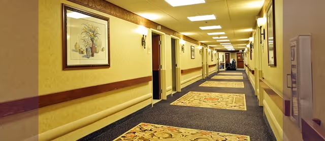 A long indoor hallway with patterned carpets and yellow walls. Several doors line the corridor on both sides, with framed artwork hanging on the walls. The ceiling has recessed lighting and wall sconces provide additional light.