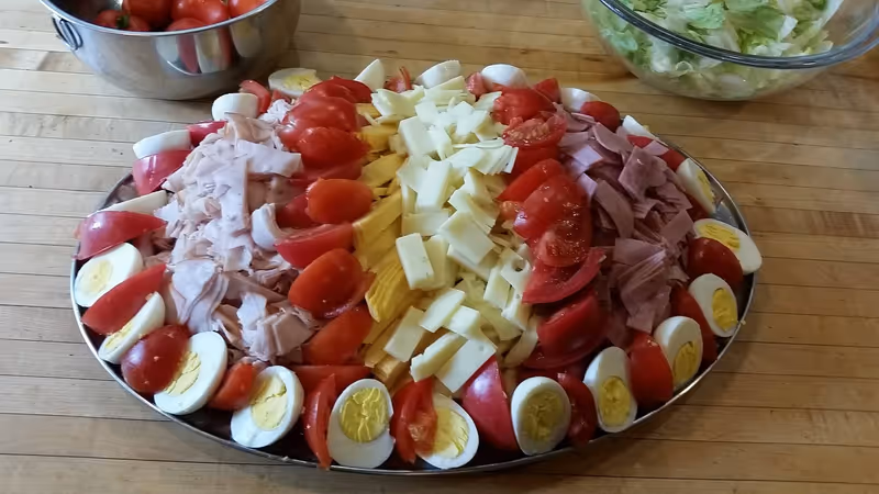 A large oval platter with neatly arranged sliced hard-boiled eggs, tomato wedges, shredded turkey, sliced cheese, and sliced ham on a wooden table. In the background, there is a metal bowl with cherry tomatoes and a glass bowl with lettuce.