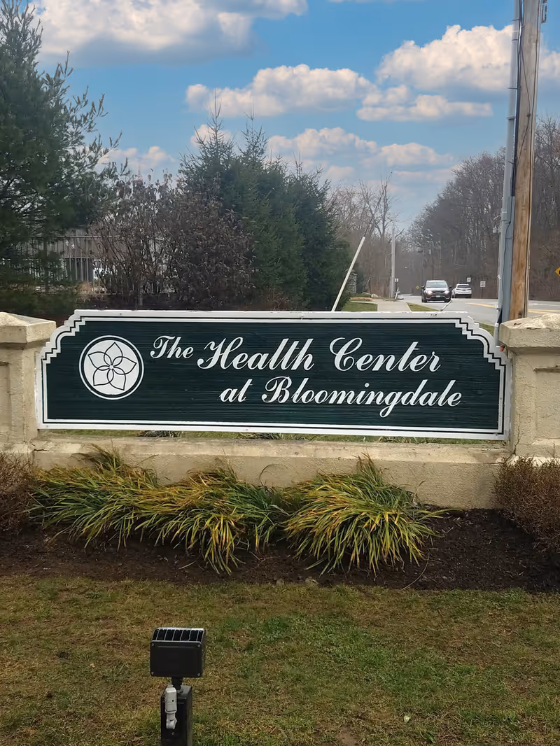 Outdoor sign for The Health Center at Bloomingdale mounted on a stone base with greenery and trees in the background under a partly cloudy sky.