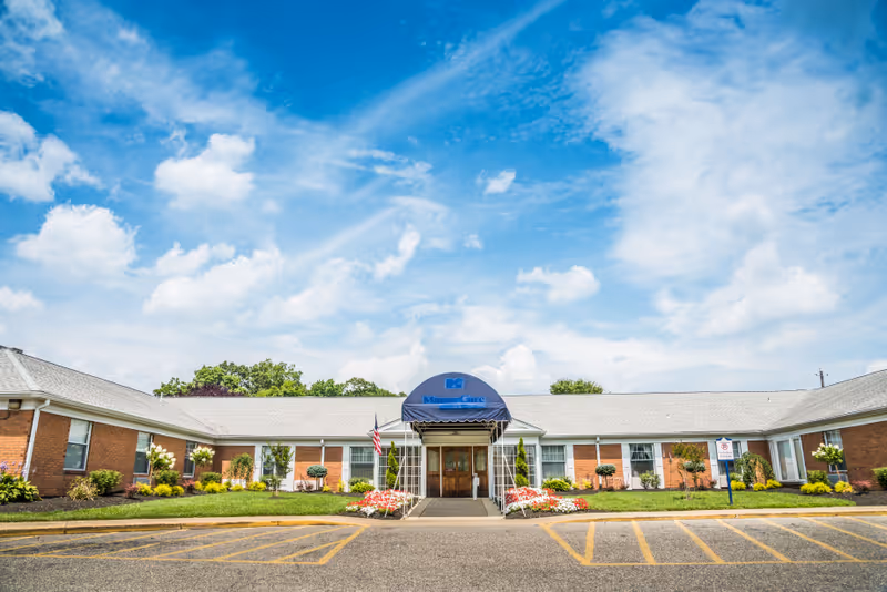 Front exterior view of Barclays Rehabilitation And Health Care Center, a single-story brick building with a gray roof, a blue awning over the entrance, landscaped greenery, flowers, and an American flag near the entrance under a partly cloudy blue sky.