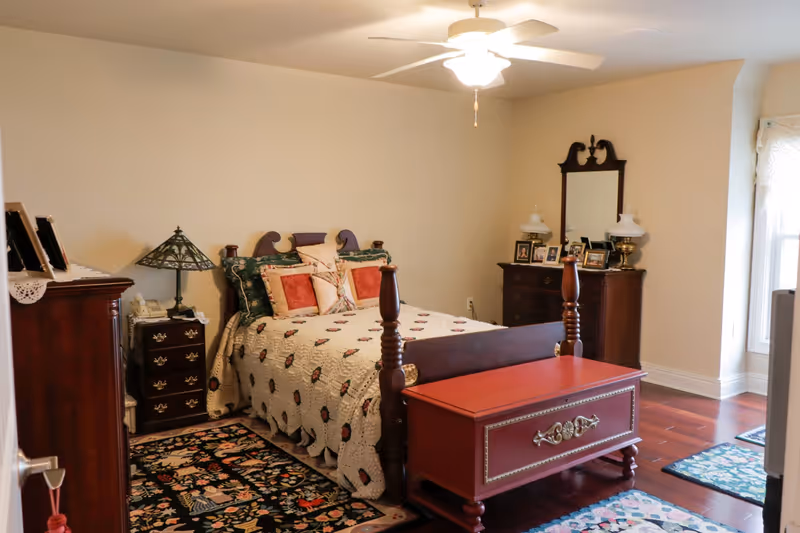 A cozy bedroom with a wooden bed frame, a quilted bedspread, and multiple pillows. There is a wooden chest at the foot of the bed, a nightstand with a decorative lamp and a telephone, a dresser with framed photos and two lamps, and a ceiling fan with a light. The room has hardwood floors and several patterned rugs.