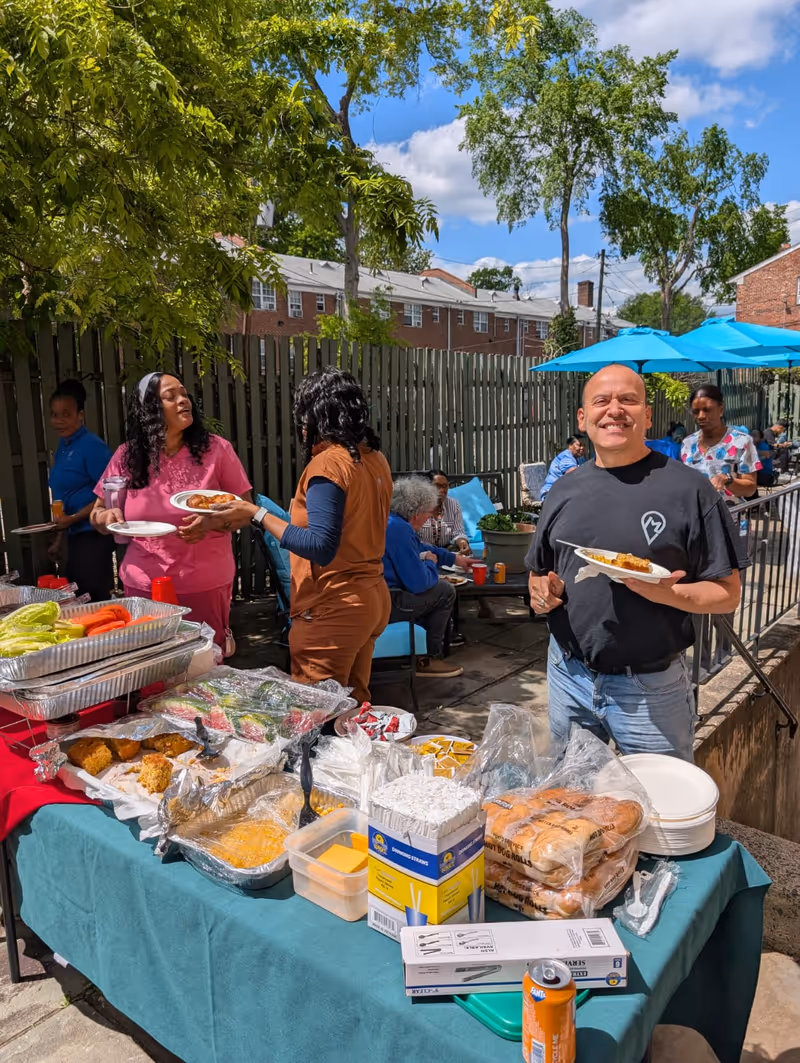 Outdoor gathering at Montclair Care Center with people serving and enjoying food at a buffet table covered with a green tablecloth. The table has trays of food, buns, cheese slices, and condiments. Several people are standing and sitting under blue umbrellas with a wooden fence and trees in the background on a sunny day.