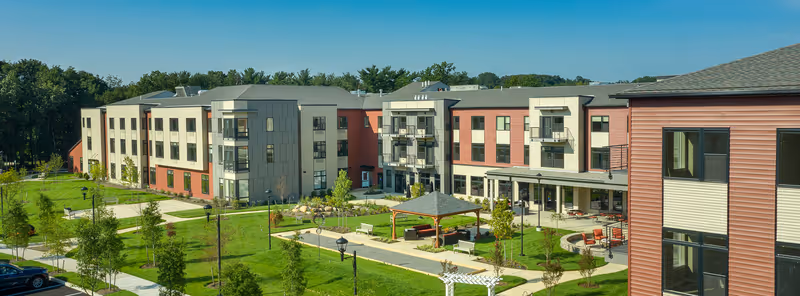 Exterior view of a senior living facility with a three-story building featuring balconies, large windows, and a landscaped courtyard with green lawns, trees, benches, walking paths, and a gazebo under a clear blue sky.