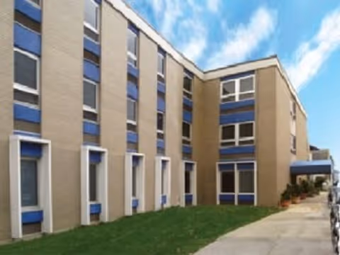 Exterior view of a multi-story building with beige and blue accents, a green lawn in front, and a sidewalk leading to an entrance under a blue sky with some clouds.