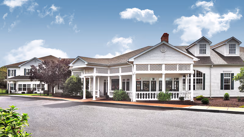 Front exterior of a white, colonial-style senior living building with a covered porch entrance, columns and a driveway.