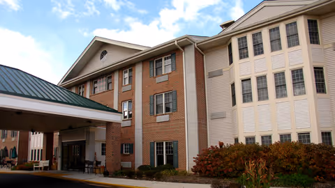 Exterior view of a multi-story senior living facility building with brick and beige siding, multiple windows, a covered entrance, and landscaping with bushes under a partly cloudy sky.