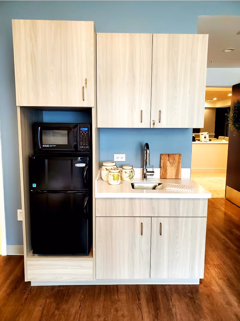 A small kitchenette area with light wood cabinets, a black microwave and mini refrigerator, a white countertop with a sink and faucet, decorative jars, and a wooden cutting board. The floor is wooden, and the wall behind the kitchenette is painted blue.