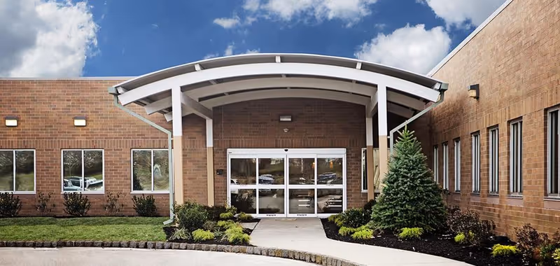 Front exterior view of a brick building with a covered entrance featuring a curved white canopy. There are several windows on both sides of the entrance, landscaped bushes, and a small evergreen tree near the walkway leading to the automatic glass doors. The sky is partly cloudy.