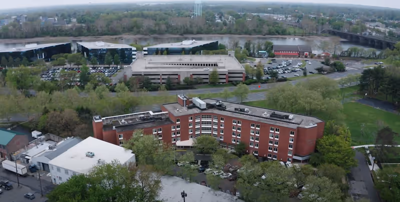 Aerial view of a multi-story red-brick rehabilitation and nursing center surrounded by trees, with a river and parking structures in the background.