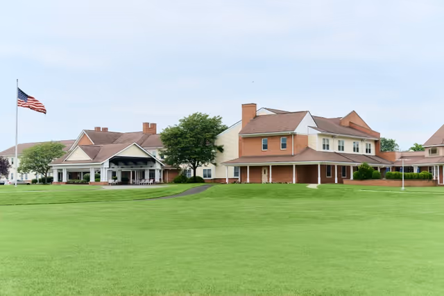 Wide view of Berlin Rehabilitation & Healthcare Center showing multiple connected buildings with brick and light-colored siding, surrounded by a large green lawn and an American flag on a flagpole.