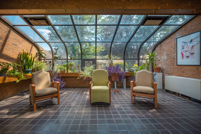 A sunroom with a glass ceiling and walls, featuring three chairs arranged in a row on a tiled floor. The room has brick walls and is decorated with various potted plants along the sides and back. A framed picture of birds hangs on the right wall.