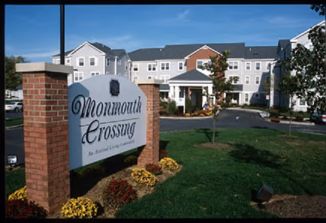 Exterior view of Monmouth Crossing, a residential living community building with a large sign in front surrounded by landscaped grass and flowers under a clear blue sky.