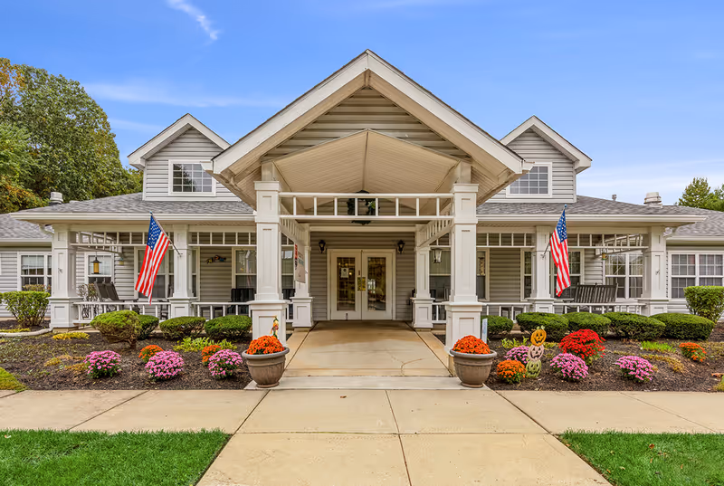 Front exterior view of Summit Place facility with a covered entrance, two American flags, well-maintained flower beds with colorful flowers, and a clear blue sky.