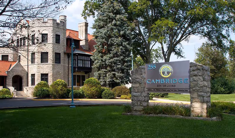 Stone building with a castle-like turret and red roof surrounded by trees and bushes, with a stone sign in front that reads '255 Cambridge Senior Living' on a sunny day.
