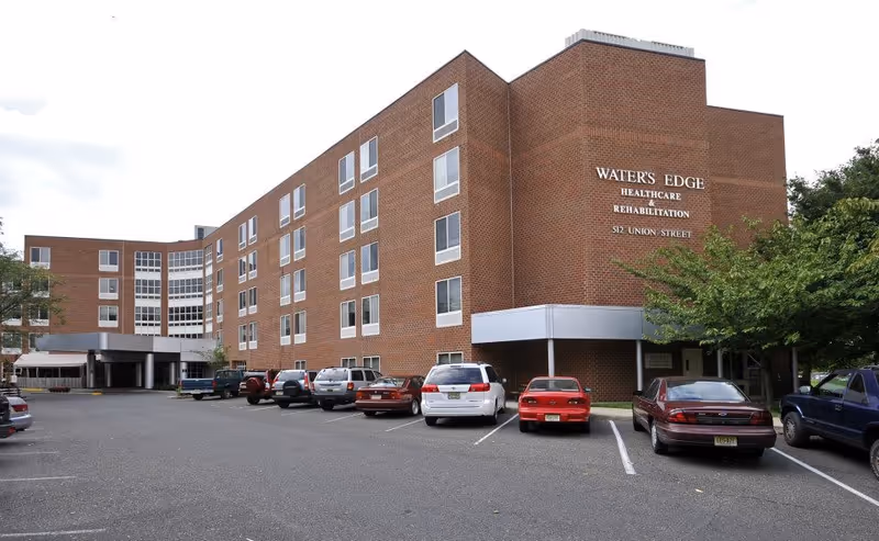Exterior view of a multi-story brick healthcare and rehabilitation building with several parked cars in front. The building has multiple windows and a covered entrance area. Trees are visible on the right side of the image.