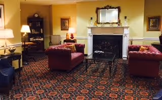 A cozy living room with two red sofas facing each other across a glass coffee table, a fireplace with a mirror above it, a wooden bookshelf, and warm lighting from table lamps. The room has patterned carpet flooring and light yellow walls with framed artwork.