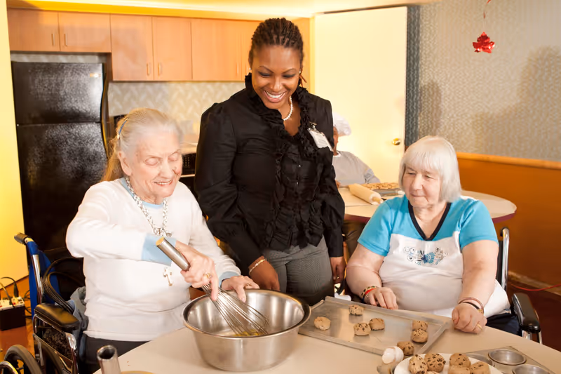 Two elderly women sitting at a table in a kitchen area, one whisking ingredients in a large metal bowl while the other looks on. A smiling caregiver stands between them. There is a tray with cookie dough and baking utensils on the table, and a man in the background wearing a chef's hat.