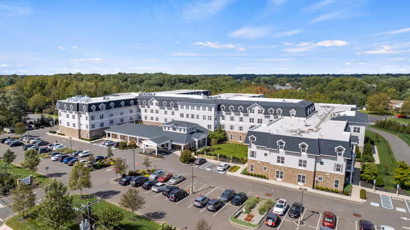 Aerial view of a large senior living facility building with multiple floors, surrounded by a parking lot filled with cars and green trees in the background under a blue sky with some clouds.