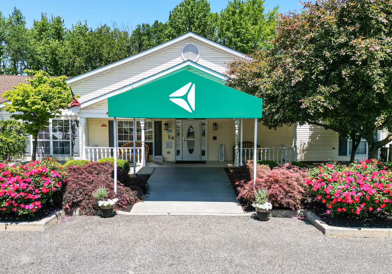 Front exterior view of Arden Courts - ProMedica Memory Care Community in Cherry Hill, showing a single-story building with a green awning over the entrance, surrounded by well-maintained landscaping including colorful flowers and trees.