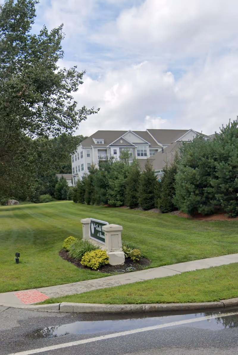 Exterior view of the Health Center at Bloomingdale showing a large, multi-story residential building surrounded by green trees and well-maintained grass. A sign with the facility's name is placed on a small landscaped area near the sidewalk and road.