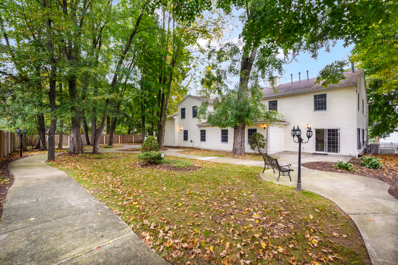 A peaceful outdoor area at Fox Trail Memory Care Living featuring a winding concrete pathway surrounded by green grass and trees with autumn leaves scattered on the ground. There is a white two-story building with multiple windows and a sliding glass door. A black metal bench and a vintage-style street lamp are positioned near the pathway.