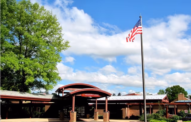 Exterior view of Meadow Lakes facility with a covered entrance, brick pillars, and an American flag flying on a flagpole under a partly cloudy blue sky.