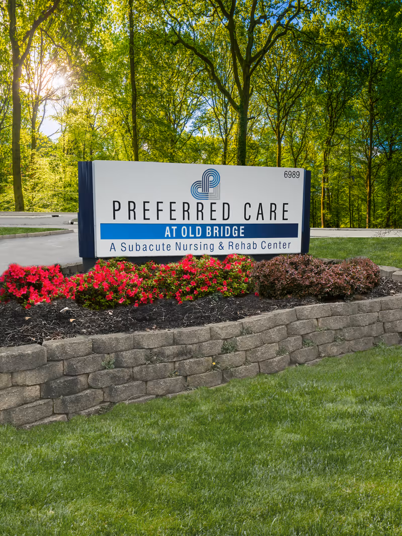 Outdoor sign for Preferred Care at Old Bridge, a subacute nursing and rehab center, surrounded by a landscaped garden with red flowers and green bushes, with trees and a road in the background.