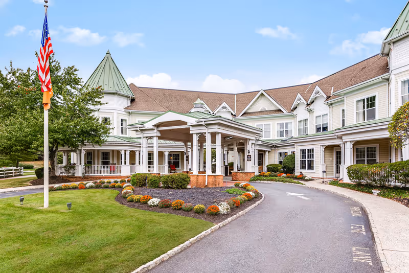 Front entrance of a senior living building with a covered porte-cochere, landscaped circular driveway, and an American flag.