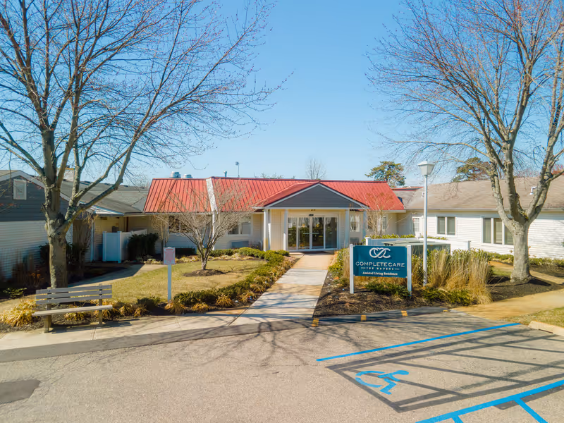 Front entrance of an assisted living building with a red metal roof, walkway, landscaped beds, bench, and a 'Complete Care' sign.