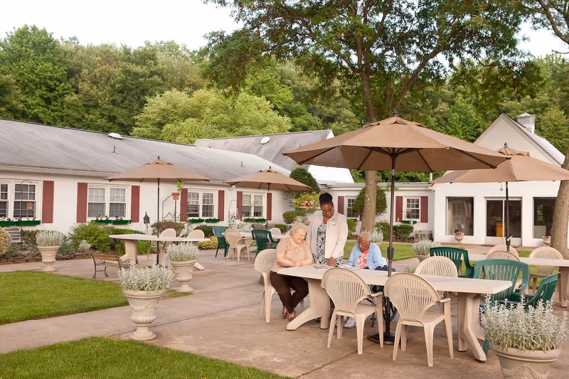 Outdoor patio area at Ashbrook Care & Rehabilitation Center with tables, chairs, and large umbrellas. Two elderly women are seated at a table, one in a wheelchair, and a staff member is standing with them, engaging in conversation. The background shows a white building with red shutters and green plants, surrounded by trees.