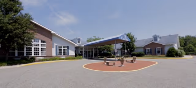 Front exterior view of CareOne at Morris facility showing a large driveway with a covered entrance, brick buildings on either side, and some greenery around the building.