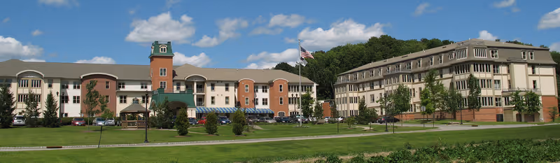 Exterior view of Bristol Glen, a UMC Community, showing two large multi-story buildings with a green lawn and trees in front under a blue sky with scattered clouds. An American flag is flying on a flagpole near the center of the image.