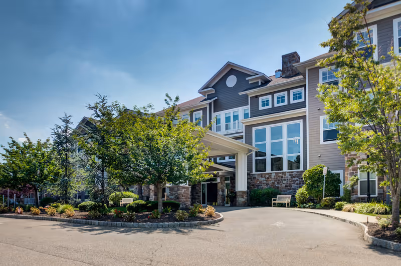 Front exterior of a multi-story senior living building with a covered entrance, landscaped trees and benches under a blue sky.