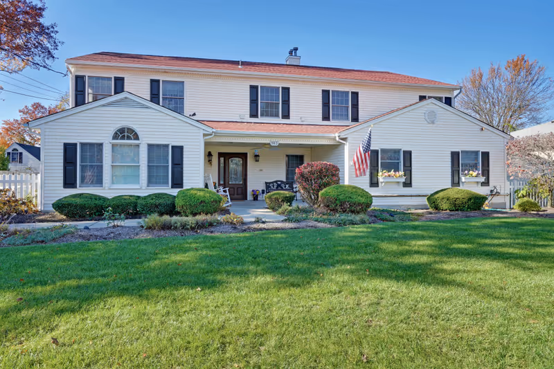 Front exterior view of a two-story white residential-style building with black shutters and a red roof, surrounded by neatly trimmed bushes and a green lawn. An American flag is displayed near the entrance, which has a covered porch with seating.