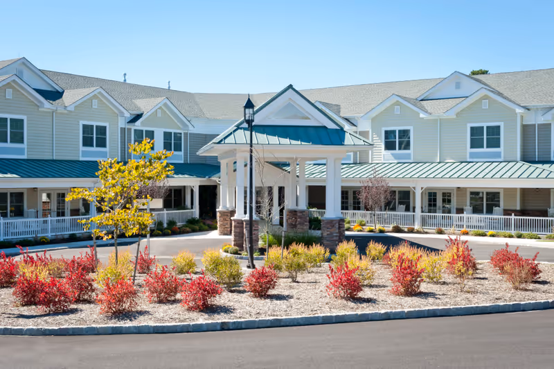 Exterior view of a senior living facility with a covered entrance supported by white columns and stone bases. The building has beige siding with white trim and a green metal roof. In front of the entrance, there is a landscaped area with small trees and red and yellow shrubs under a clear blue sky.