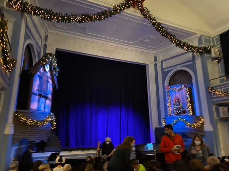 An indoor stage decorated with Christmas garlands and lights, featuring a large blue curtain. People wearing masks are gathered in front of the stage, some standing and some seated. The room has ornate architectural details and festive holiday decorations.