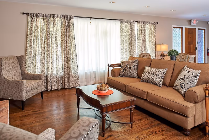 A cozy living room with a brown sofa adorned with patterned cushions, two upholstered armchairs with a subtle pattern, a wooden coffee table with decorative items, patterned curtains covering large windows, and a wooden floor. A lamp and a potted plant are visible near the entrance door in the background.